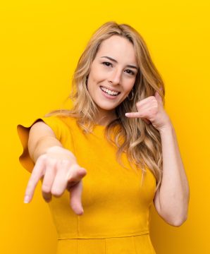 young pretty blonde woman smiling cheerfully and pointing to camera while making a call you later gesture, talking on phone against flat color wall
