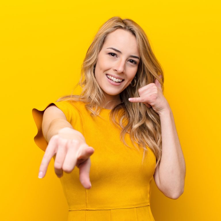 young pretty blonde woman smiling cheerfully and pointing to camera while making a call you later gesture, talking on phone against flat color wall