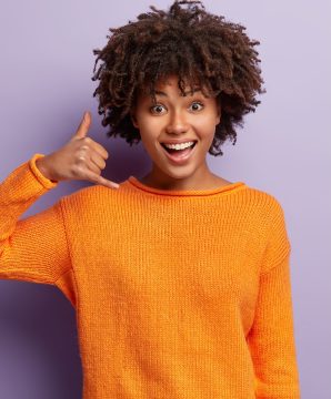 Delighted young female model with happy expression, makes call gesture, asks being in touch, wears casual orange jumper, isolated over purple background with free space for your advertisement