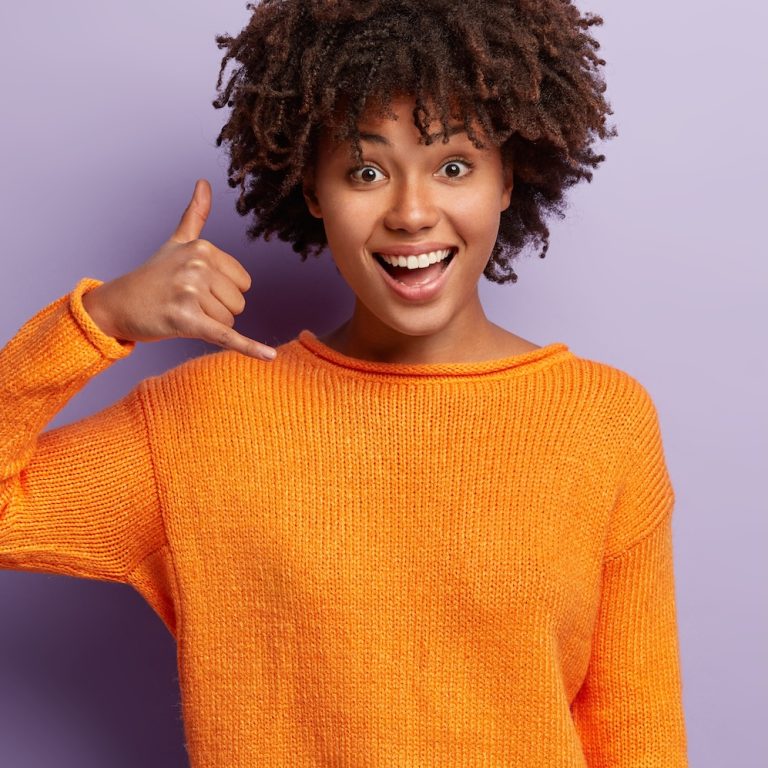 Delighted young female model with happy expression, makes call gesture, asks being in touch, wears casual orange jumper, isolated over purple background with free space for your advertisement