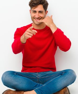 young handsome man smiling cheerfully and pointing to camera while making a call you later gesture, talking on phone sitting on the floor