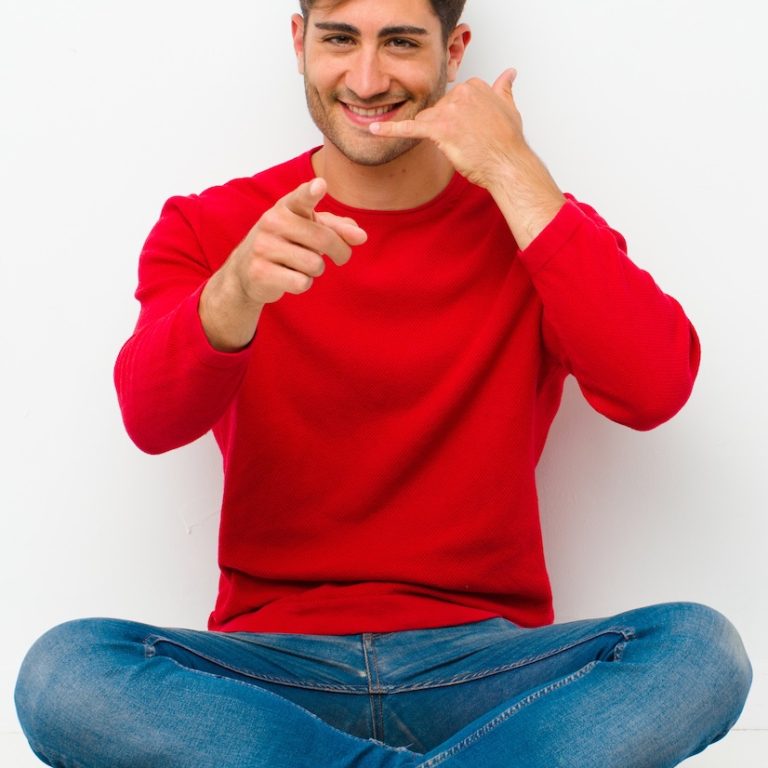 young handsome man smiling cheerfully and pointing to camera while making a call you later gesture, talking on phone sitting on the floor