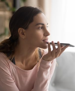 Woman sitting on couch holds phone talks on speakerphone with friend, makes voice recognition or request uses internet services through virtual assistant, record audio message, translator app concept
