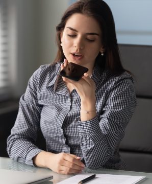 Young confident female manager worker entrepreneur using mobile voice recognition app, searching information in internet at workplace. Friendly businesswoman recording voice message on smartphone.