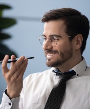 Head shot close up happy millennial businessman holding smartphone near mouth, using virtual assistant service. Smiling successful entrepreneur recording voice message in messenger on mobile phone.