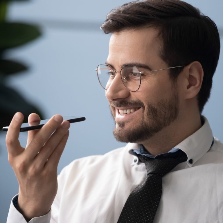 Head shot close up happy millennial businessman holding smartphone near mouth, using virtual assistant service. Smiling successful entrepreneur recording voice message in messenger on mobile phone.