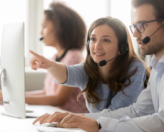 Female call center agent operator telemarketer helping male colleague teaching new worker online customer support pointing at computer, business team in wireless headset talk working together on pc